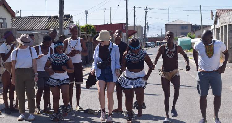 Touristen und lokale Tanzende, die gemeinsam auf einer Straße in einem Township auftreten, die von einfachen Häusern gesäumt ist.