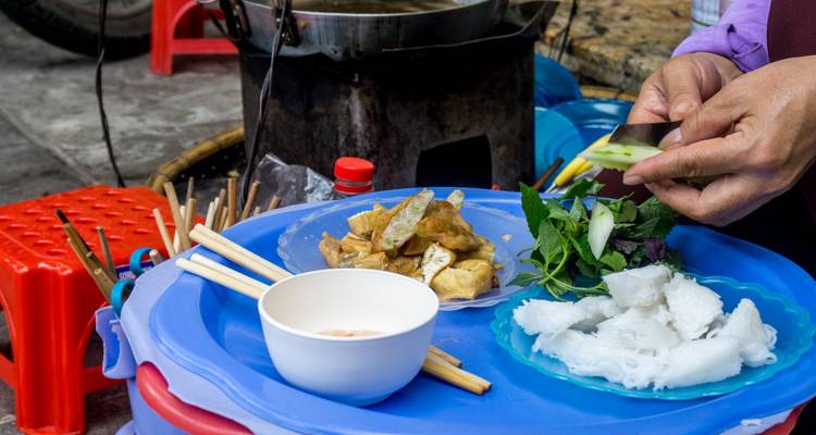 Street-food vendor preparing tofu, herbs and rice noodles on a blue plastic table.