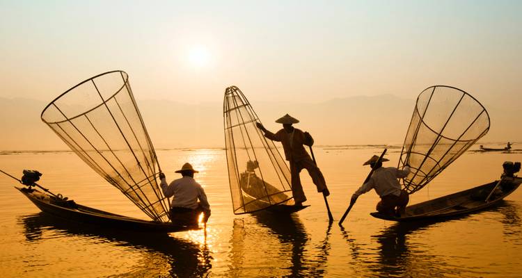 Silhouetted fishermen balancing on narrow boats with conical nets at sunrise over calm water.