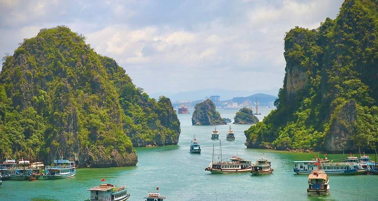 Numerous tourist boats navigating between towering limestone karsts in Halong Bay under partly cloudy skies.