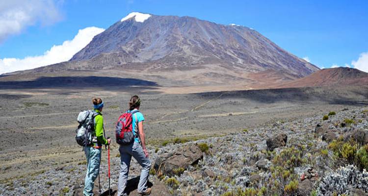Twee wandelaars pauzeren op een vulkanisch plateau en bewonderen de enorme kegel van de Mount Kilimanjaro die voor hen uitrijst.