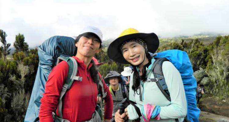 Twee vrouwelijke trekksters met grote rugzakken die lachen op een bosrijk pad van de Mount Kilimanjaro.