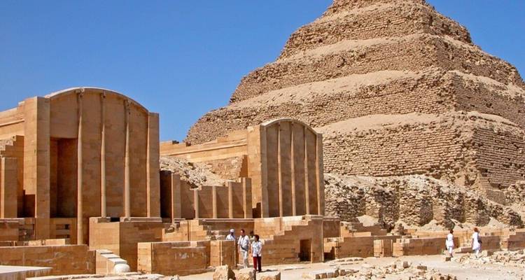 La Pirámide Escalonada de Saqqara se alza en capas de piedra caliza detrás de las ruinas de un templo antiguo bajo un cielo impoluto.