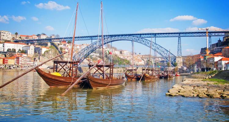 Traditional wooden rabelo boats float on the Douro River with the Dom Luís I Bridge and Porto skyline behind.