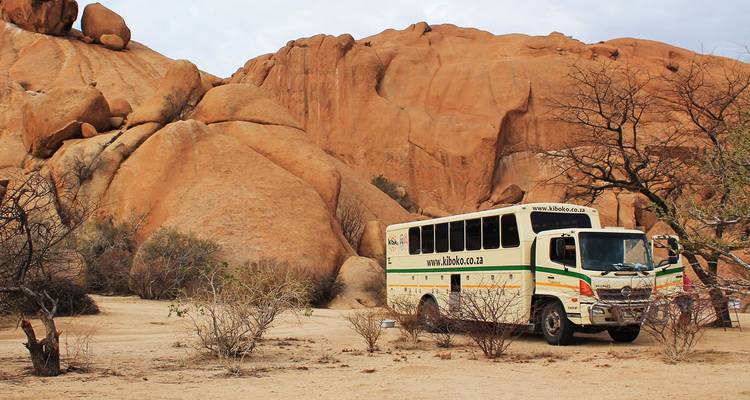 Autobus de safari terrestre garé parmi d'énormes rochers de granit orange et une végétation désertique clairsemée.
