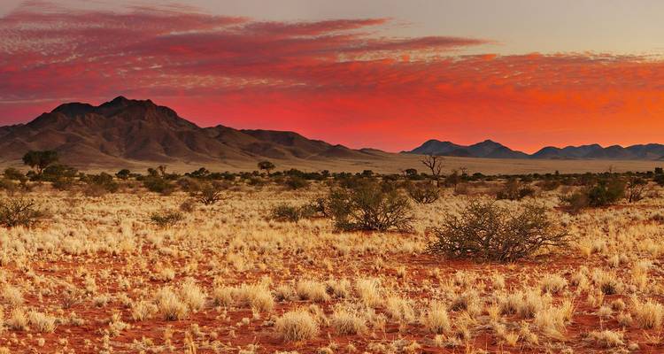 Cielo ardiente al atardecer sobre pastizales secos y montañas lejanas en el desierto de Namib.