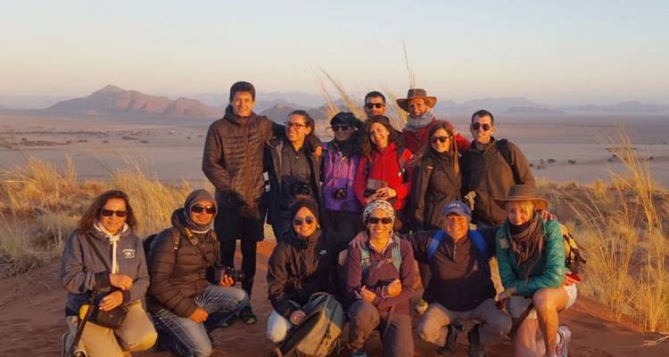 Grupo de viajeros posando en una duna al amanecer con llanuras desérticas y montañas de fondo.