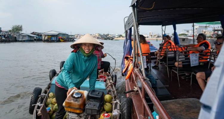 Un vendeur souriant portant un chapeau conique vend des noix de coco depuis un petit bateau motorisé le long d'un bateau de tourisme sur une rivière.