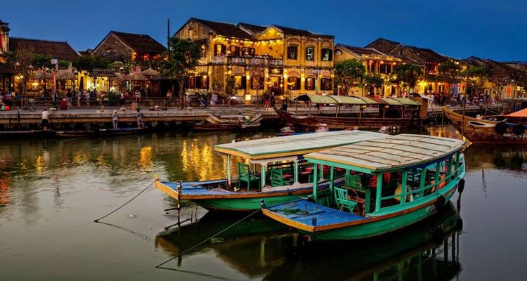 Des bateaux en bois illuminés par des lanternes amarrés sur le fleuve devant la façade fluviale brillante de Hoi An au crépuscule.