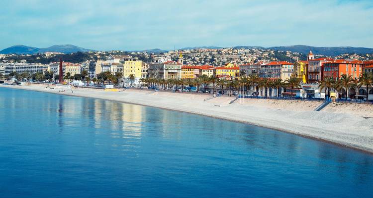 Longue plage de galets et promenade bordée de palmiers adossée à des bâtiments colorés à Nice sur la Côte d'Azur.