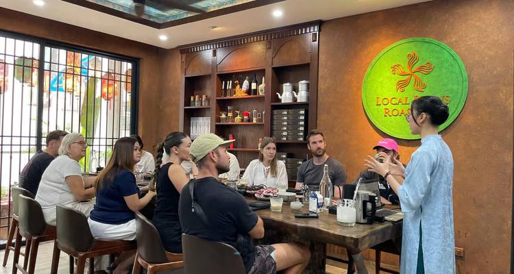 Taller de café donde viajeros sentados alrededor de una mesa de madera escuchan la presentación de un barista