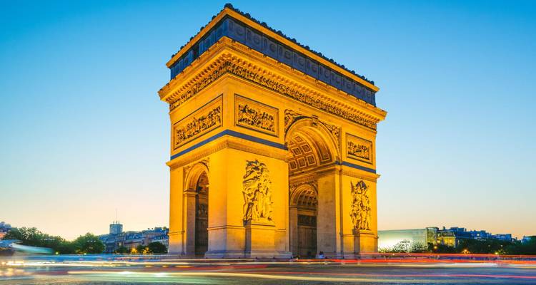 El Arco de Triunfo iluminado con una luz dorada contra un cielo crepuscular despejado con rastros de tráfico coloridos rodeándolo.