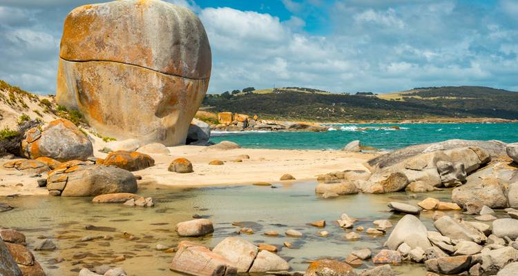 Gran roca de granito redondeada en una playa de arena con piscinas poco profundas y un mar turquesa al fondo.