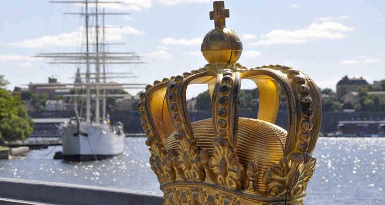 Detalle de una corona dorada ornamentada en el Puente Skeppsholmsbron con un barco de mástil alto desenfocado al fondo.