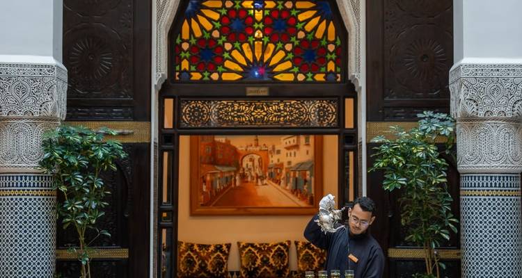 Un serveur dans un riad traditionnel verse le thé à la menthe sous une arche de vitrail colorée.