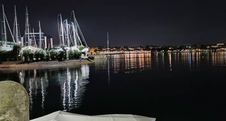 Vista nocturna de un puerto junto al lago con mástiles de veleros y luces del pueblo reflejándose en el agua