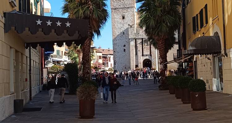 Torre de piedra medieval y calle bulliciosa en Malcesine con multitudes bajo un cielo azul brillante