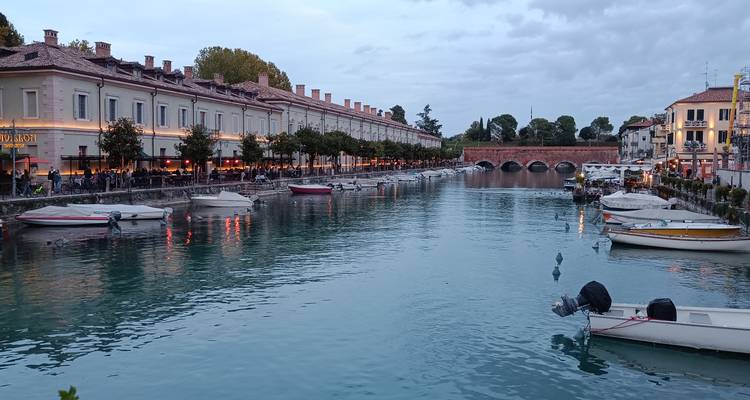 Vista nocturna del canal de Peschiera del Garda con barcos amarrados a los lados y restaurantes iluminados, un puente de piedra al fondo