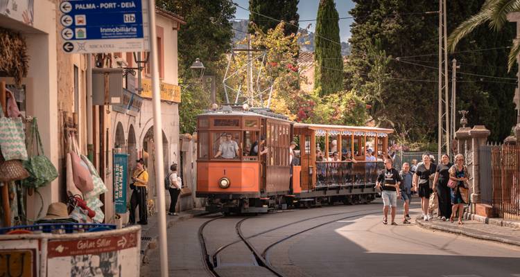 Tramway vintage en bois orange rempli de passagers tournant dans une rue bordée de fleurs dans un village historique.