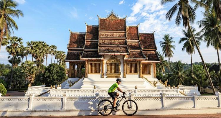 Cycliste en maillot vert passant devant un temple laotien orné avec des palmiers sous un ciel bleu.
