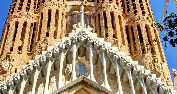 Vue de face de la façade de la Passion de la Sagrada Família avec des colonnes angulaires et des lettres gravées sous un ciel ensoleillé.