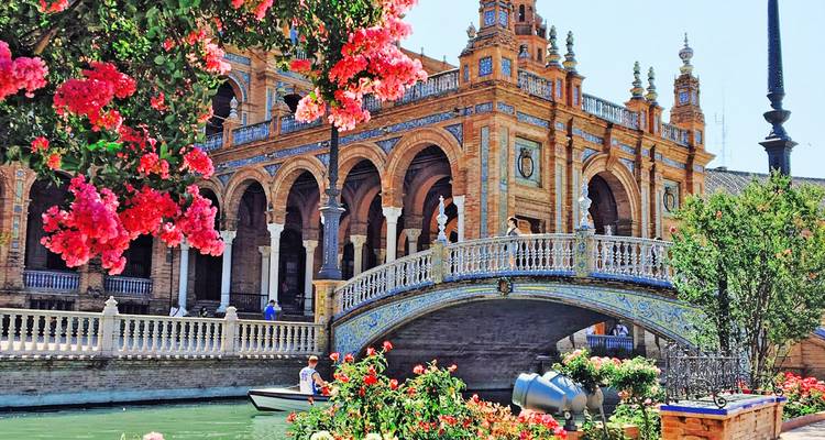 Las flores coloridas enmarcan el puente ornamentado, los arcos y el canal de la Plaza de España de Sevilla con un bote de remos abajo.