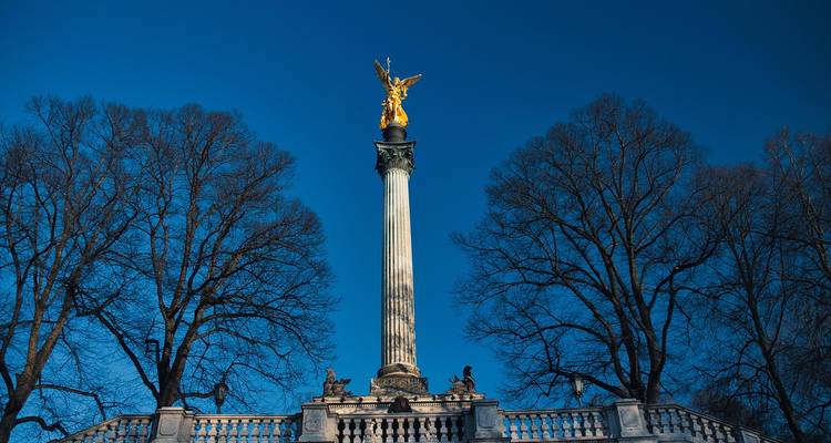 Estatua de ángel dorado en lo alto de la columna blanca Friedensengel rodeada de árboles sin hojas contra un cielo azul profundo en Múnich