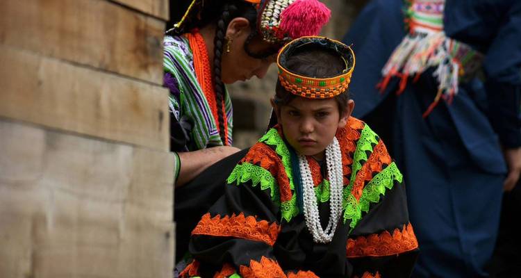 Junges Kalash-Kind in aufwendigem orange- und grünfarbenem Kostüm, das nachdenklich schaut, während ein Ältester die Kleidung dahinter anpasst.