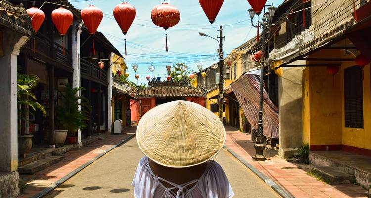 Achteraanzicht van een persoon in een bamboe punthoed die onder rode lantaarns door een oud straatje in Hoi An loopt.