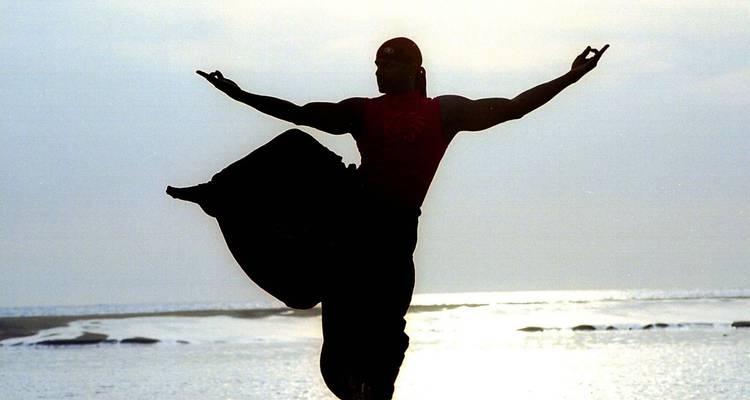 Silhouet van een martial artist die een dynamische pose aanneemt op een strand bij zonsopgang met de glinsterende zee op de achtergrond.
