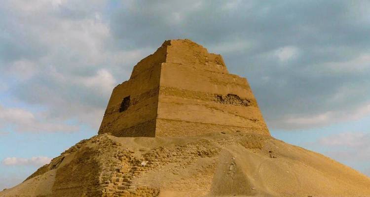 Meidum-stijl trappenpiramide die zich verheft uit een zandheuveltje onder een bewolkte hemel