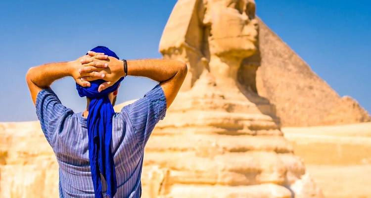 Een jonge reiziger staat met zijn handen achter zijn hoofd geklemd en kijkt naar de Sphinx en een piramide tegen een heldere blauwe lucht.
