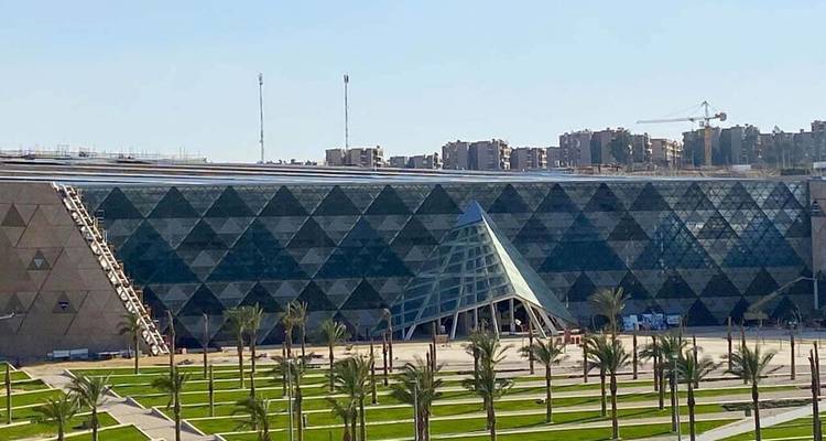 Buitenkant van het moderne Grand Egyptian Museum met een geometrisch glazen gevel en een plein omzoomd met palmen.