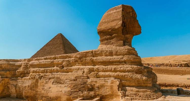 Zijkant van de Sphinx die naar een piramide kijkt onder een strakblauwe hemel.