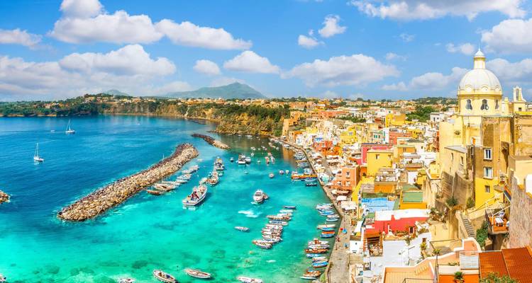 Levendige pastelkleurige gebouwen liggen langs een glinsterende turquoise haven vol met boten onder verspreide wolken in Procida.