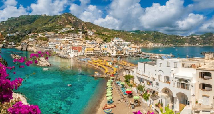 Uitzichtpunt omgeven door bloemen met zicht op een halvemaanvormig strand en kleurrijke heuveldorpje aan de kust van Ischia.