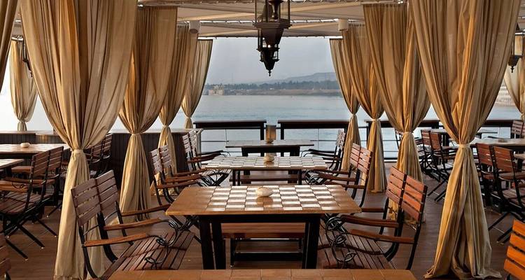 Terrasse de restaurant élégante en plein air avec tables en bois, rideaux en toile et vue sur le Nil