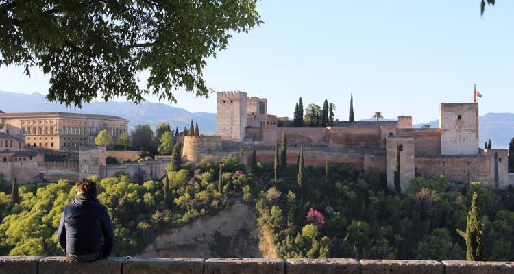 Een reiziger zit op een stenen muur en geniet van het panoramische uitzicht op het Alhambra aan de overkant van een bosrijke kloof.