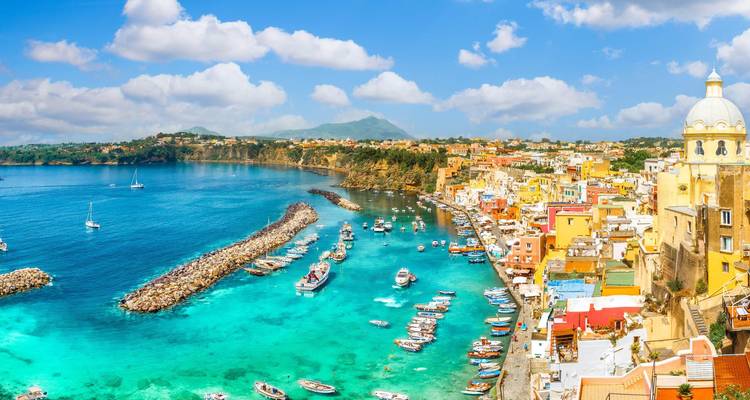 Las aguas turquesas brillantes y las casas de colores pastel de la isla de Procida se curvan alrededor de un rompeolas rocoso bajo nubes esponjosas.