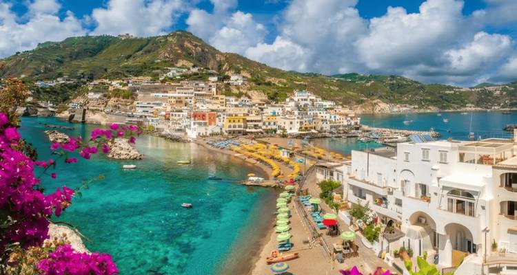 Pueblito costero encantador en Ischia con casitas de colores, sombrillas de playa y agua turquesa rodeada de buganvilias moradas.