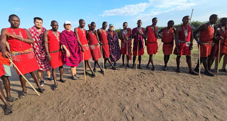 Fila de guerreros Maasai en shukas rojos parados con turistas en terreno polvoriento bajo un cielo brillante.