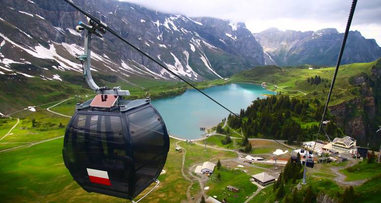 Cabina de teleférico deslizándose sobre un lago alpino rodeado de prados verdes y cimas nevadas en Suiza.
