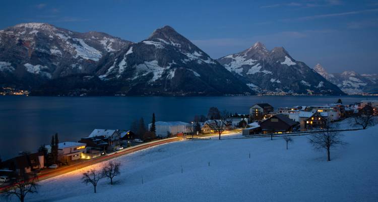 Pueblito suizo cubierto de nieve junto a un lago, enmarcado por montañas imponentes y sendas de luces de autos iluminadas al atardecer.
