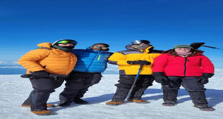 Quatre alpinistes vêtus de vestes colorées se tenant debout au sommet enneigé avec un ciel bleu profond en arrière-plan.