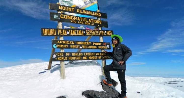 Alpiniste vêtu d'une veste sombre et d'un bonnet vert se tenant fièrement à côté du panneau du sommet du Kilimandjaro sur un sol enneigé.