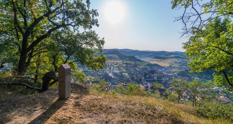 Panoramablick auf ein Talstädtchen mit einer befestigten Kirche unter strahlender Sonne, umrahmt von bewaldeten Hängen und einem Wegweiser aus Stein.