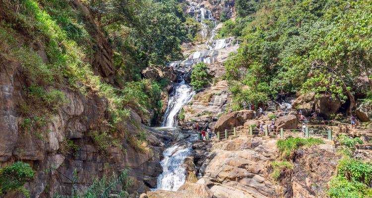 Une haute cascade se déverse à travers des falaises rocheuses dans un bassin entouré d'une forêt luxuriante avec des visiteurs sur un pont.