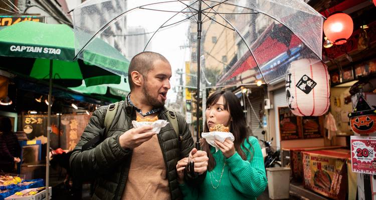 Couple mangeant de la nourriture de rue sous un parapluie transparent dans une rue de marché animée et pluvieuse.