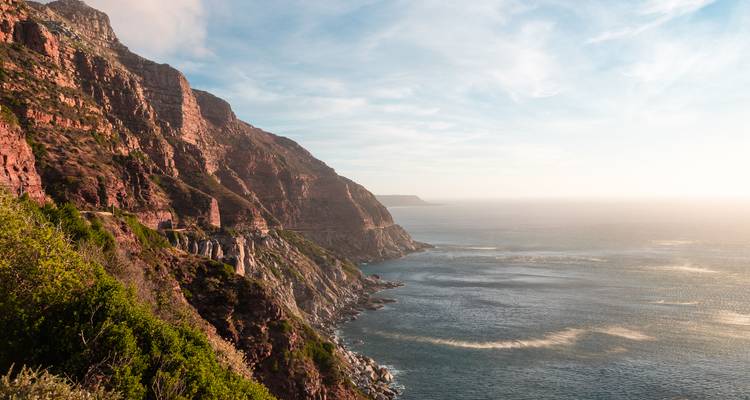 Rugged red-cliff coastline along Chapman’s Peak Drive meeting the calm Atlantic Ocean under light clouds.