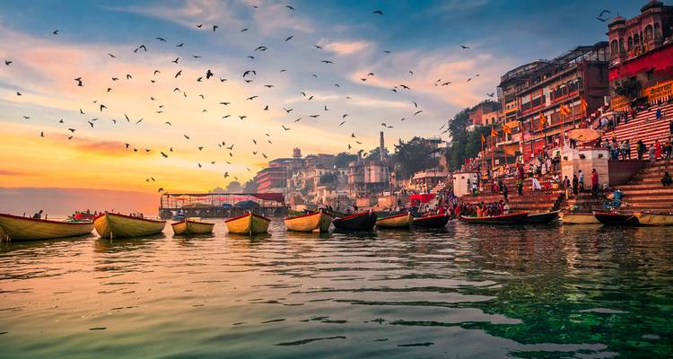 Colorful boats lined along the Ganges ghats with birds flying across a vivid sunset sky